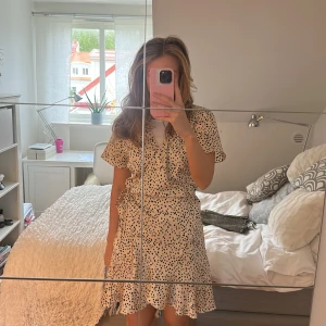 Prickig kortklänning från Neo Noir - Supersöt kortklänning från Neo Noir i beige med svarta prickar. Klänningen har korta ärmar, volangdetalj nertill och en lätt v-ringning. Perfekt för sommarens alla tillfällen!