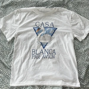 Vit t-shirt med svan och tryck från Casa Blanca - Vit t-shirt med tryck av en svan och blått rutmönster på bröstet. Texten 'Casa Blanca Par Avion' i blått framtill. Klassisk passform och rund halsringning. Perfekt för en stilren och chill look.
