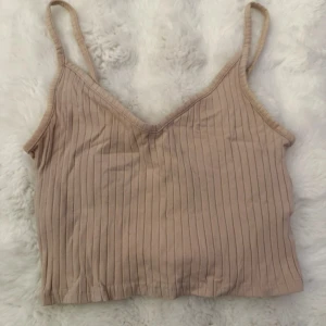 Beige ribbad linnetopp från Divided - Säljer en beige ribbad linnetopp från Divided med smala axelband och v-ringning. Toppen är croppad och har en mjuk, stretchig känsla. Perfekt att matcha med jeans eller kjol.