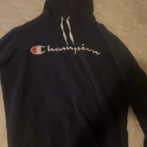 Mörkblå hoodie från Champion - Säljer en mörkblå hoodie från Champion med vit logga och snören i halsen. Klassisk modell med huva och mjukt material, perfekt för chill dagar eller när du vill vara extra bekväm.
