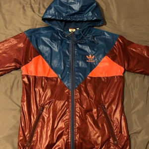 Adidas vindjacka med huva - Säljer en retroinspirerad Adidas vindjacka i glansigt material. Jackan är vinröd med blå och orange detaljer, har huva och dragkedja framtill samt två fickor. Stort Adidas-tryck på ryggen och klassisk logga på bröstet.