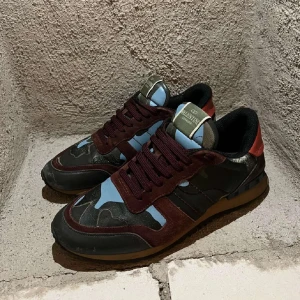 Valentino Rockrunner sneakers  - Säljer ett par Valentino Rockrunner sneakers med unikt blått och svart kamouflagemönster, vinröda detaljer och röd häl. Skorna har snörning, rund tå och platt sula. Perfekta för dig som vill sticka ut med exklusiva och trendiga sneakers.