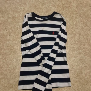 Randig långärmad tröja från Ralph Lauren - Säljer en klassisk långärmad tröja från Ralph Lauren med breda marinblå och vita ränder. Tröjan har rund halsringning och en röd broderad logga på bröstet. Perfekt för en stilren och tidlös look.Skriv till mig om ni har nån fråga eller nått om tröjan tröjan  eller om storlek för jag vet inte exakt vad storleken på tröjan är!