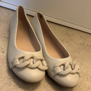 Beiga ballerinaskor med kedjedetalj - Snygga beiga ballerinaskor med en chunky kedjedetalj framtill. Skorna har rund tå och platt sula. Använda en gång. Passar 39/40