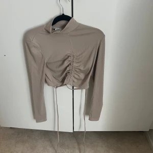 Beige croppad polotopp med snörning - Trendig beige croppad polotopp med långa ärmar och snygg snörningsdetalj framtill. Toppen har en ribbad struktur och är perfekt för att skapa en cool och stilren look.