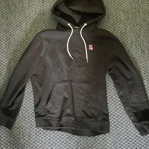 Svart hoodie från Bondelid - Svart hoodie från Bondelid med vit dragsko och en liten röd broderad detalj på bröstet. Klassisk känguruficka framtill och mjuk insida. Perfekt för en avslappnad stil.