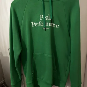 Peak Performance hoodie - Säljer en grön hoodie från Peak Performance med vit broderad logga på bröstet och känguruficka framtill. Tröjan har huva med dragsko och långa ärmar. Perfekt för en avslappnad stil.