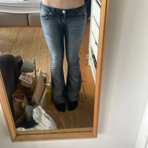 Blå bootcut jeans med låg midja - Säljer ett par blå bootcut True religion jeans med låg midja och klassiska fickor bak med dekorativa sömmar. Jeansen har en lätt tvättad look och stängs med dragkedja och knapp. Perfekta för dig som gillar en retro vibe. Aldrig använda 