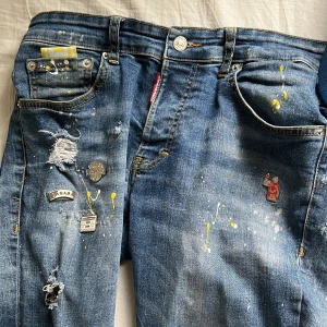 Blå jeans från dsquared2 - Unika blå jeans med flera pins, slitningar och färgstänk i gult och vitt. Byxorna har raka ben och klassisk femficksdesign. Snygga detaljer som trasiga partier och broderad patch på ena benet. Perfekta för dig som vill sticka ut med din stil.