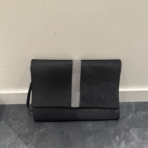 Svart kuvertväska med glittrig detalj från Atmosphere - Säljer en svart kuvertväska från Atmosphere med en snygg glittrig remsa framtill. Väskan har en elegant struktur och stängs med magnetknapp. Perfekt för att lyfta din outfit med lite extra bling. Har aldrig använt den så väskan är i nyskick! 