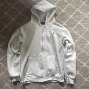  Ralph Lauren zip tröja - Säljer en klassisk vit hoodie från Polo Ralph Lauren med dragkedja och huva. Tröjan har två fickor framtill och den ikoniska broderade loggan på bröstet. Perfekt för en stilren och avslappnad look.
