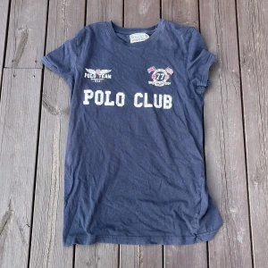 Mörkblå t-shirt Saint Vincent Polo - Mörkblå t-shirt från Saint Vincent med tryck 'POLO CLUB' och coola emblem framtill. Klassisk rund hals och korta ärmar. Skön passform och tillverkad i mjuk bomull, perfekt för en avslappnad stil.