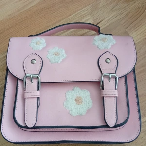 Rosa handväska med blommor - Supergullig ljusrosa handväska med broderade vita blommor på locket. Väskan har ett klassiskt skolväskesnitt med två spännen framtill och ett handtag upptill. Tillverkad i syntetmaterial och har svarta kanter som kontrast.