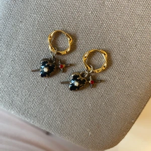 Guldörhängen med svarta hjärtan och svärd🫶 - Unika örhängen med tvinnade guldfärgade ringar och hängande svarta hjärtan dekorerade med vita detaljer. Genom hjärtat går ett litet svärd med röd sten, vilket ger en edgy vibe. Perfekt för dig som vill sticka ut med coola accessoarer.🍁🍁