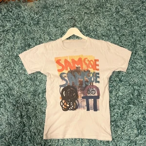 Vit t-shirt med färggrafiskt tryck Samsoe - Vit t-shirt från Samsoe Samsoe i storlek S med ett stort, färgglatt grafiskt tryck på framsidan. Trycket har inslag av orange, blått, gult och svart med coola illustrationer och text. Klassisk rund hals och korta ärmar. Perfekt för dig som gillar streetstyle och unika prints.