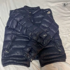 Mörkblå dunjacka från Moncler - Säljer en mörkblå dunjacka från Moncler med klassisk quiltad design och hög krage. Jackan har dragkedjor på fickorna och tryckknappar vid ärmsluten. Perfekt för kalla dagar och riktigt snygg till streetwear-stilen. Tyvärr nåt hål på ärmarna och svår och knäppa