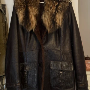Brun skinnjacka med päls från Alviero Armani - Säljer en brun lång skinnjacka från Alviero Armani med stor fluffig päls på kragen. Jackan har två stora fickor med flätad detalj och är helfodrad med mjuk päls på insidan. Perfekt för dig som vill sticka ut med en lyxig och vintage-inspirerad look.