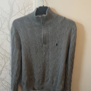 Grå stickad tröja Polo Ralph Lauren - Kabelstickad grå tröja från Polo Ralph Lauren med half zip och hög krage. Klassisk broderad logga på bröstet och ribbade muddar. Perfekt för lager-på-lager och snygg till jeans eller chinos.
