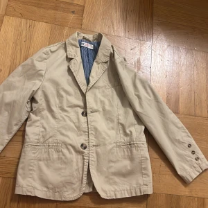 Beige kavaj från H&M, strl 128 - Snygg beige kavaj från H&M i storlek 128. Klassisk modell med två knappar framtill, fickor och krage. Insidan har ett blåvitt randigt foder som ger en cool kontrast. Perfekt för dig som gillar stilrena och enkla plagg.
