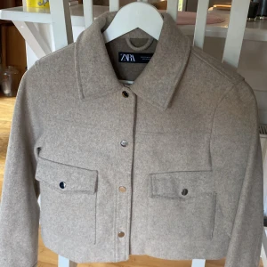 Beige overshirt från Zara i ullmix - Säljer en beige overshirt från Zara i ullmix med klassisk krage, stora bröstfickor med lock och knappar framtill. Jackan är croppad och har en clean, stilren look som passar perfekt till lager-på-lager. Långärmad och med en mjuk känsla.