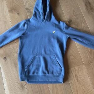 Blå hoodie från Lyle & Scott  - Snygg blå hoodie från Lyle & Scott Junior med klassisk gul logga på bröstet. Tröjan har huva, känguruficka och långa ärmar. Skick: 9,5/10✅ Passar cirka 158cm.