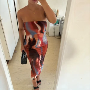 Tubklänning med abstrakt mönster - Säljer en snygg axelbandslös tubklänning i midi-längd med abstrakt mönster i rött, blått, vitt och brunt. Klänningen är figurnära och har en tight passform, perfekt för dig som vill sticka ut. Materialet känns stretchigt och följsamt.