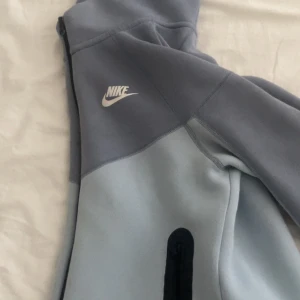 Nike tech hoodie i två nyanser av blått - Nike tech hoodie i två olika blå nyanser med vit logga på bröstet. Tröjan har huva, lång ärm och en svart dragkedja på sidan. Perfekt för dig som gillar sportig och avslappnad stil.