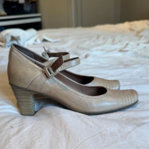 Beige Mary Jane-klackar från Tamaris - Snygga Mary Jane-klackar från Tamaris i beige skinn med rund tå och dekorativa sömmar framtill. Skorna har en stabil låg klack och justerbart spänne över foten. Perfekt för dig som gillar klassisk stil med en modern twist.