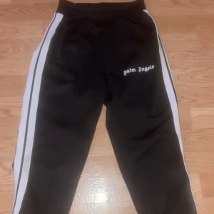 Palm Angels svarta byxor  - Svarta mjukisbyxor från Palm Angels med vita sidoränder och broderad logga på låret. Byxorna har elastisk midja med snörning och är tillverkade i ett mjukt, syntetiskt material. Perfekta för en chill och sportig look.Och har 2 fickor vid sidan,kommer med tags