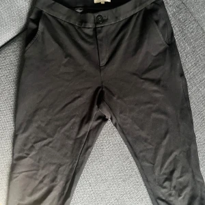 Svarta kostymbyxor Part Two strl 34 - Svarta kostymbyxor från Part Two i klassisk rak modell med fickor i sidorna och knappstängning. Byxorna har uppvikta benslut och en stilren look som passar till många olika outfits. Materialet känns mjukt och följsamt.