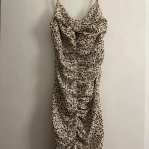 Blommig omlottklänning med volang - Supersöt kort klänning med blommigt mönster i beige, brunt och grönt. Klänningen har omlottdetalj vid bysten, tunna axelband och snygg volang framtill. Perfekt för sommarens alla tillfällen och ger en fräsch och trendig vibe.