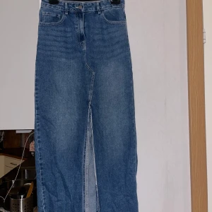 Lång jeanskjol med slits och rå kant - Snygg lång jeanskjol i klassisk blå denim med hög midja, slits framtill och rå, fransig kant nertill. Kjolen har femficksmodell och stängs med knapp och dragkedja. Perfekt för en chill och trendig look.