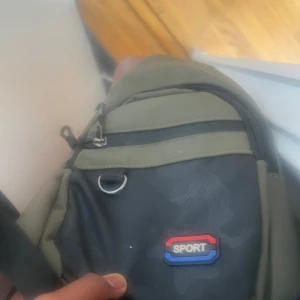 Grön och svart ryggsäck SPORT - Säljer en stilren ryggsäck i grönt och svart med SPORT-logga framtill. Väskan har flera dragkedjefack, en metallring för nycklar eller accessoarer och justerbara axelband. Perfekt för skola eller träning och rymmer allt du behöver till vardagen.