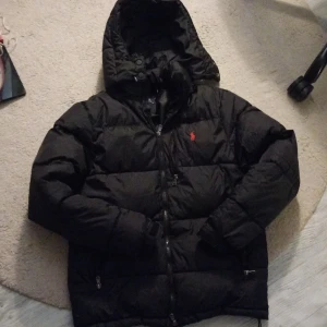 Svart dunjacka från Polo Ralph Lauren - Säljer en svart dunjacka från Polo Ralph Lauren med huva och dragkedja. Jackan har en klassisk pufferstil och är försedd med Polo-loggan broderad i rött på bröstet. Perfekt för kalla dagar och riktigt snygg till streetwear-looken.