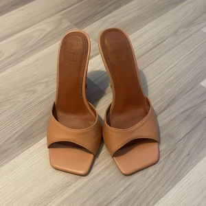 Beige klackade mules med fyrkantig tå - Snygga beige mules från Guess med öppen häl och fyrkantig tå. Skorna har hög klack och bred rem över foten. Tillverkade i slätt skinnliknande material, perfekta för att lyfta din outfit med en trendig touch.