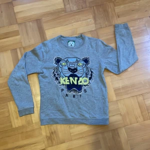 Grå Kenzo sweatshirt med tigertryck - Säljer en grå sweatshirt från Kenzo med ikoniskt broderat tigerhuvud och Kenzo Paris-logga i gult och blått framtill. Tröjan har rund hals, långa ärmar och är gjord i mjuk bomull. Perfekt för dig som vill sticka ut med en streetstyle-vibe.