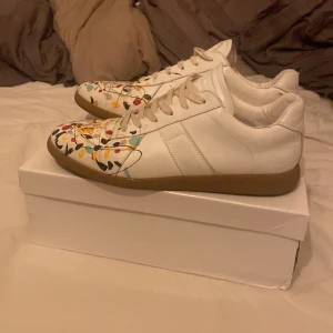 Maison Margiela sneakers med färgstänk - Unika vita sneakers från Maison Margiela med färgstänk i gult, svart, rött och turkost. Skorna har klassisk låg siluett, snörning och gummisula. Tillverkade i skinn och har en konstnärlig vibe som sticker ut. Perfekta för dig som vill ha något annorlunda. Kan tänkas gå ner i pris vid en snabb affär👍. Kolla gärna in mina andra annonser 😄