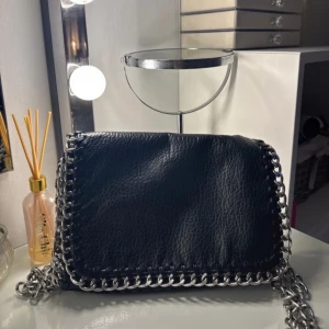 Svart axelväska med kedjedetaljer från Tiamo - Cool svart axelväska från Tiamo i skinnimitation med grova silverfärgade kedjor längs kanterna och som axelrem. Väskan har magnetlås, dragkedjefack både inuti och utanpå. 