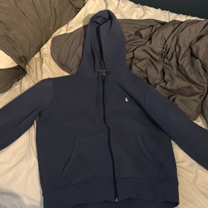 Äkta Marinblå hoodie från Polo Ralph Lauren  - Snygg marinblå hoodie från Polo Ralph Lauren med dragkedja och klassisk broderad logga på bröstet. Tröjan har huva med snörning och två stora fickor framtill. Perfekt för en chill och stilren look. Nypris runt 2500 kr