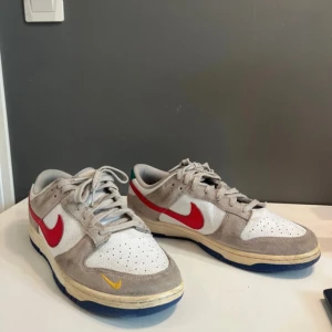 Nike Dunk Low med röd swoosh - Nike Dunk Low sneakers med vit mesh och grå mocka, röd swoosh på sidorna och liten gul swoosh framtill. Blå sula och grönt Nike-broderi på hälen. Klassisk low-top siluett med snörning, perfekt för dig som gillar streetwear.