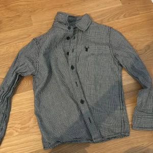 Rutig svartvit skjorta med knappar - Snygg rutig skjorta i svart och vitt med klassisk krage och långa ärmar. Skjortan har knäppning framtill och ett litet broderat emblem på bröstet. Perfekt för dig som gillar stilrena och tidlösa plagg.