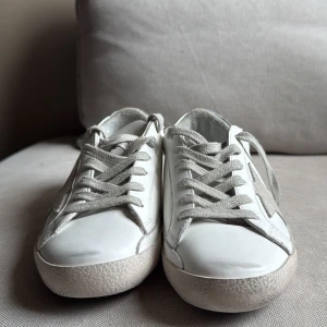 Golden Goose vita sneakers med silverdetaljer - Golden Goose sneakers i vitt skinn med grå mockadetaljer och silverfärgad häl. Klassisk stjärna på sidan, grå snörning och platt sula. Snygg och avslappnad modell som passar 37 38