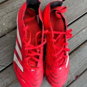 Adidas Predator röda fotbollsskor - Säljer ett par röda Adidas Predator fotbollsskor med vita och svarta detaljer. Skorna har snörning och är tillverkade i syntetmaterial. De har klassiska tre ränder på sidan och dobbar under sulan för bra grepp på planen.