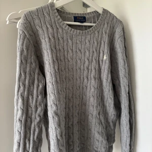 Grå kabelstickad tröja Polo Ralph Lauren - Kabelstickad grå tröja från Polo Ralph Lauren i klassisk modell. Den passar som M Tröjan har rund halsringning, långa ärmar och diskret broderad logga på bröstet. Perfekt för lager-på-lager och chill stil. Materialet är mjukt och känns riktigt skönt mot huden.