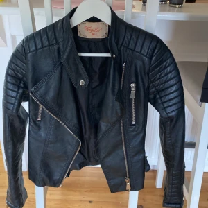 Svart skinnjacka från Voyelles, stl 36 - Snygg svart skinnjacka från Voyelles i bikerstil med coola quiltade detaljer på axlar och ärmar. Jackan har flera dragkedjor framtill, bältesdetaljer i midjan och markerad krage. Perfekt för dig som vill ha en edgy look.