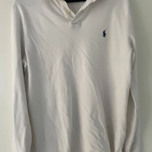 Vit långärmad pikétröja från Polo Ralph Lauren - Säljer en klassisk vit långärmad pikétröja från Polo Ralph Lauren med blå broderad logga på bröstet. Tröjan har krage och knappar vid halsen, perfekt för en clean och stilren look. 