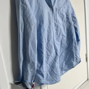 Ljusblå skjorta från Burberry - Snygg ljusblå skjorta från Burberry i bomull med klassiskt rutigt tyg på insidan av ärmsluten. Skjortan har bröstficka med broderad logga och vita knappar framtill. Perfekt för dig som gillar stilrena plagg med en twist. Använd 1-3 gånger. Säljer skjortan pga att den var för liten för mig. Storleken är s men passar även xs. Finns en liten vit fläck på baksidan av skjortan därav priset. Finns inga andra defekter på skjortan.😃🤩