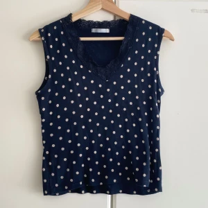 Svart prickig väst topp med spets - Supersöt svart linnetopp med vita prickar och v-ringning dekorerad med spets. Ärmlös modell som är perfekt för varma dagar. Tidlös och lekfull design som passar till jeans eller kjol.