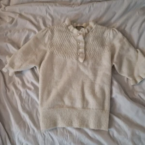 Vit stickad tröja Holly & Whyte - Supersöt vit stickad tröja från Holly & Whyte med korta puffärmar, ribbade muddar och dekorativa knappar framtill. Tröjan har ett diskret mönster upptill och en liten volangkant vid halsen. Perfekt för lager-på-lager under kyliga dagar.