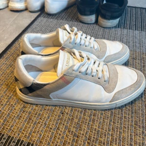 Burberry sneakers med rutigt mönster - Snygga sneakers från Burberry i vitt skinn och grå mocka med klassiskt rutigt mönster på sidorna. Skorna har snörning och rund tå, samt diskret Burberry-logga på plösen. Perfekta för dig som vill ha en stilren men ändå unik look. Pris sitter inte fast i sten😁